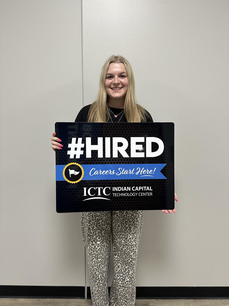 A woman holds a black sign with white text that says "#HIRED Careers Start Here!" against a white wall.