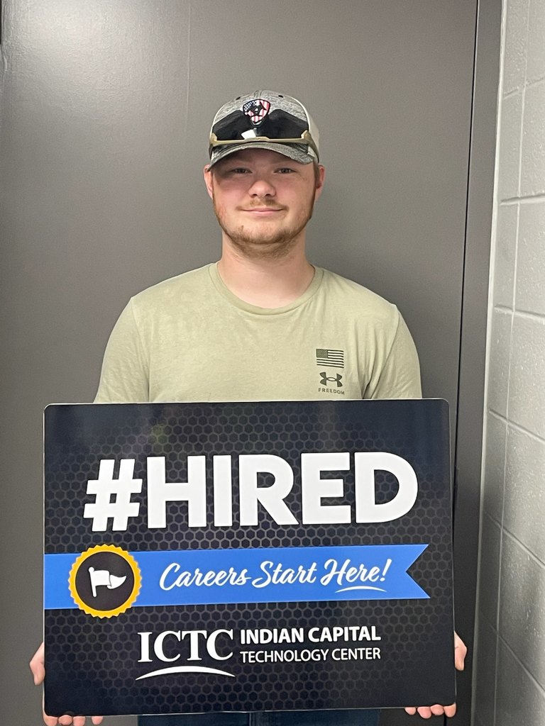A man with a hat holds a sign that says "#HIRED Careers Start Here!" in front of a gray background.