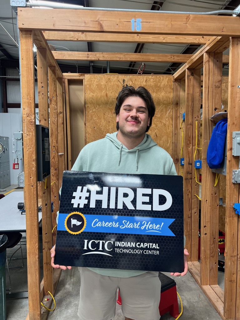 Person in a room with wooden walls, holding a sign that reads "#HIRED Careers Start Here!" and a logo.
