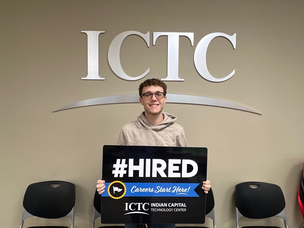 Man holds sign that reads "#HIRED" in front of a banner that says "ICTC" on a wall.