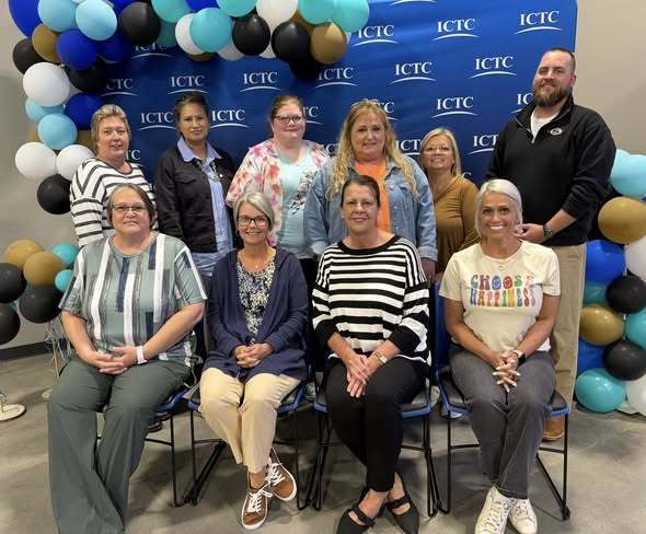 A group of people seated in chairs against a backdrop of blue and gold balloons.