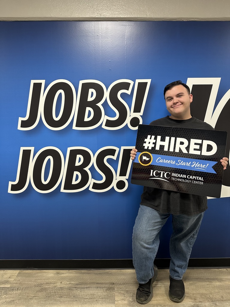 A man holds a sign that says "#HIRED" in front of a blue wall with "JOBS!" written on it.