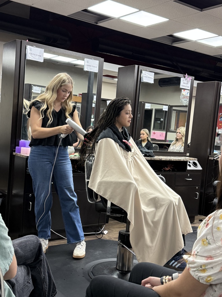 A woman in a salon styles another woman's hair with a hairdryer. Two other people sit in chairs.