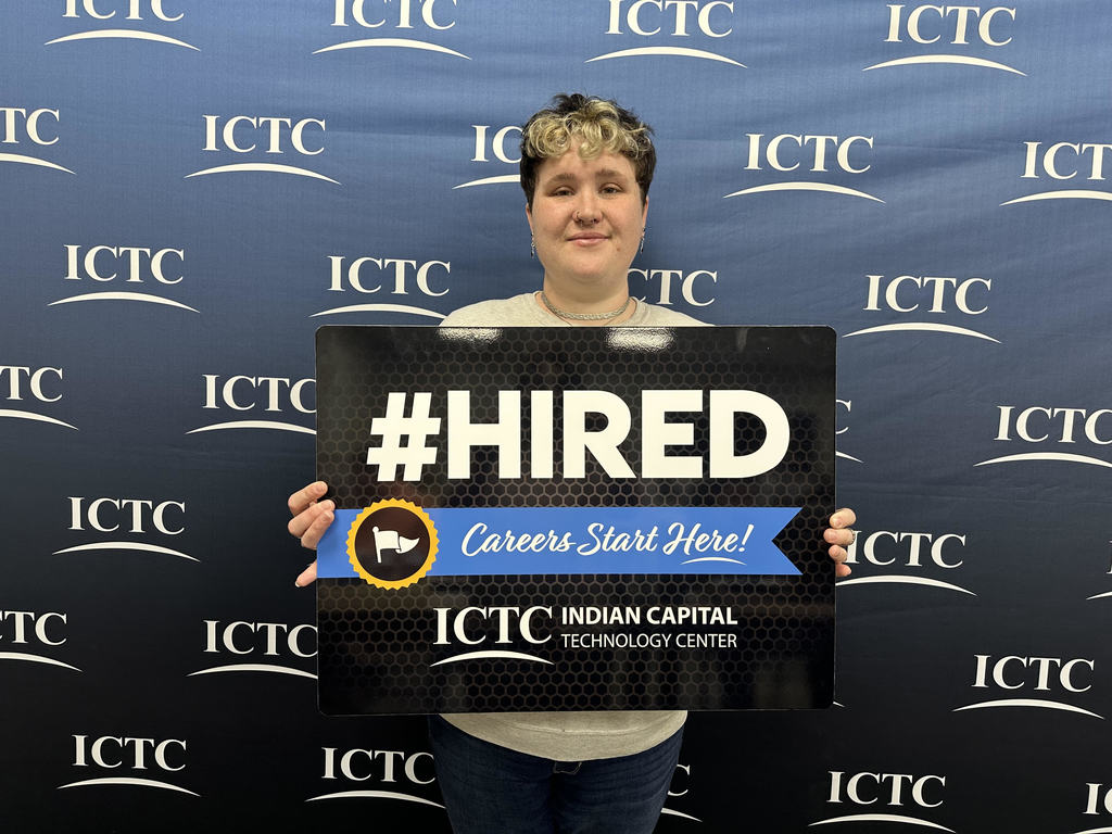 Person in front of a blue banner, holding a sign that reads "#HIRED" and "Careers Start Here!"