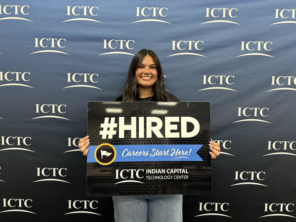A woman stands against a blue backdrop, smiling and holding a sign that reads "#HIRED" and "Careers Start Here!"