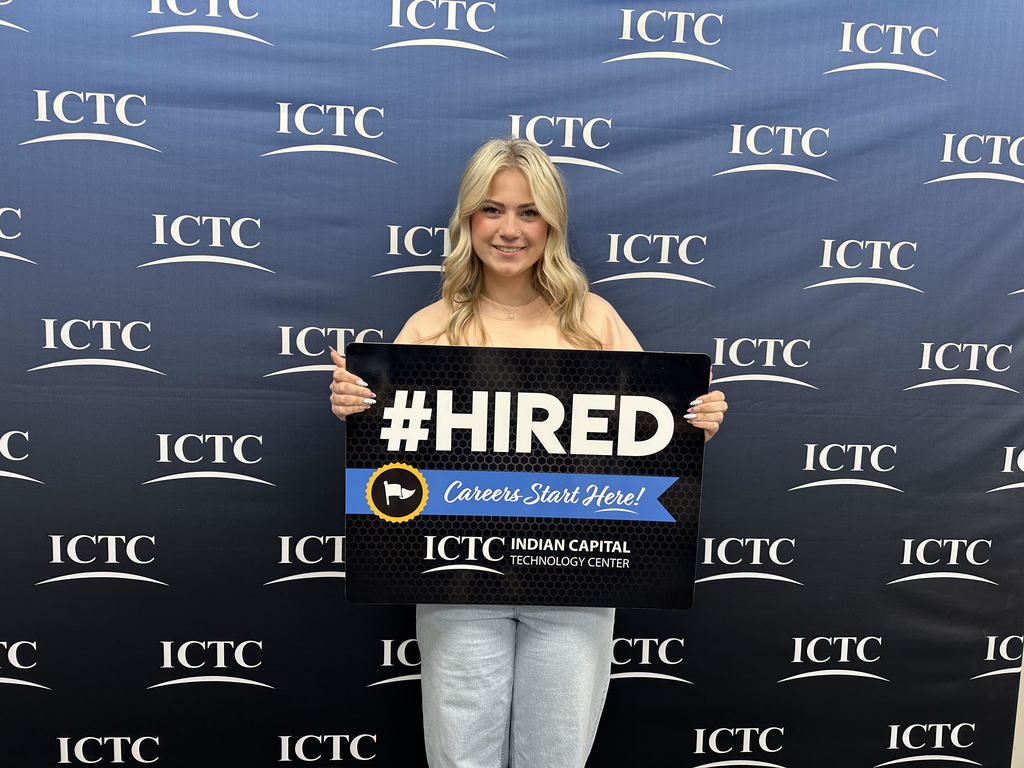 Person holding a black sign with "#HIRED" and "Career Start Here!" in front of a blue banner.