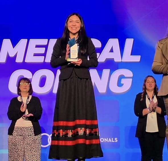 Woman holding a trophy stands on a stage with two women clapping behind her. Text behind them reads Medical Coding.