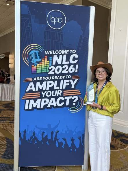 Woman in hat and glasses holding award next to a sign that says "Welcome to NLC 2026! Are you ready to amplify your impact?"