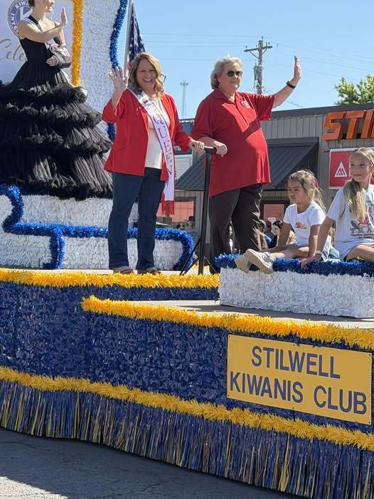 People stand on a float with "Stilwell Kiwanis Club" sign. Two women wave. Two girls sit. A woman in black dress behind them.