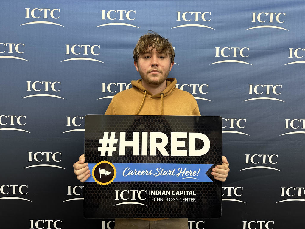 A person stands holding a black sign reading "#HIRED" against a backdrop with repeated "ICTC" branding.