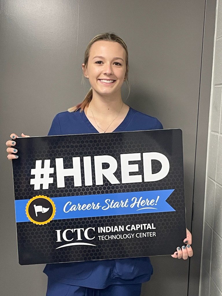 Woman in blue scrubs holds a sign with "#HIRED" and "Careers Start Here!" written on it.