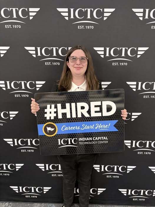 A woman poses with a sign that says "#HIRED" in front of a black background with white logos.