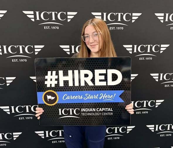 A woman holds a "#HIRED" sign against a backdrop with logos and text including "ICT." She wears glasses and smiles.