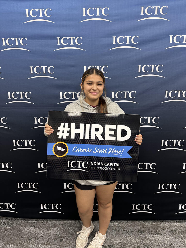 Woman holding a sign with "#HIRED" and "Careers Start Here!" in front of a blue banner.