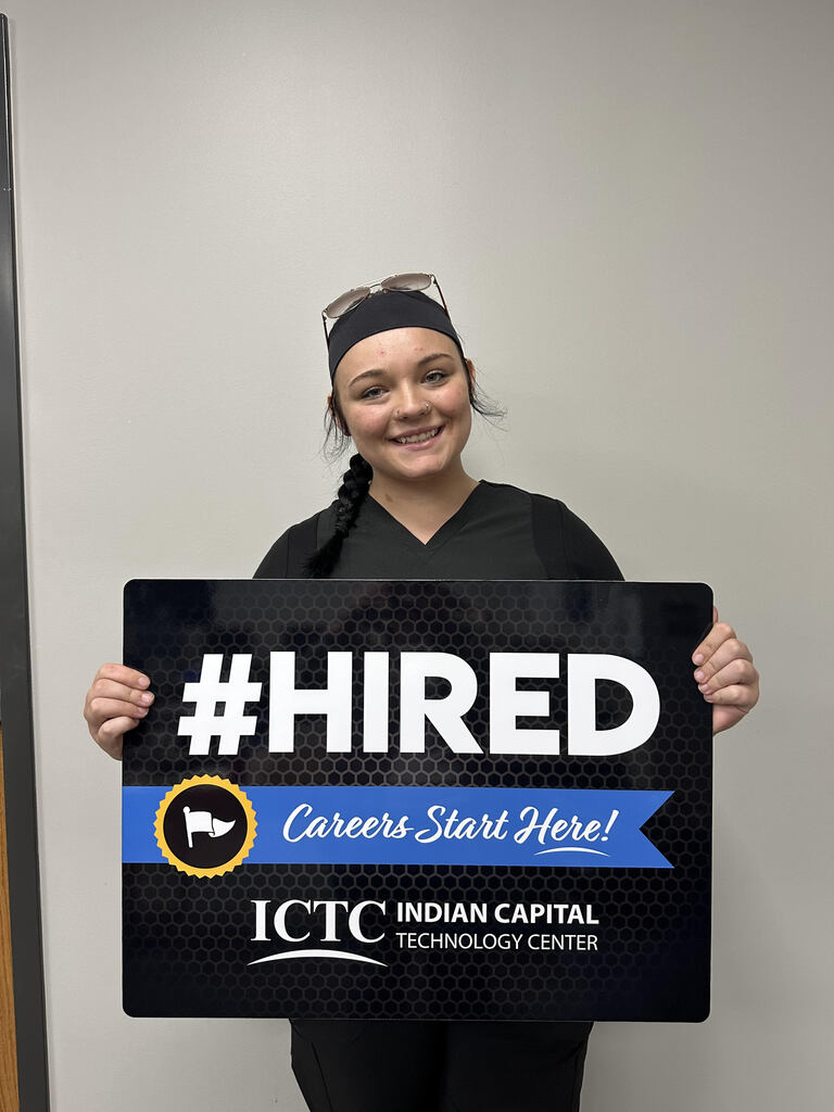 A smiling woman holds a black sign with "#HIRED" in white, a blue ribbon, and logo.