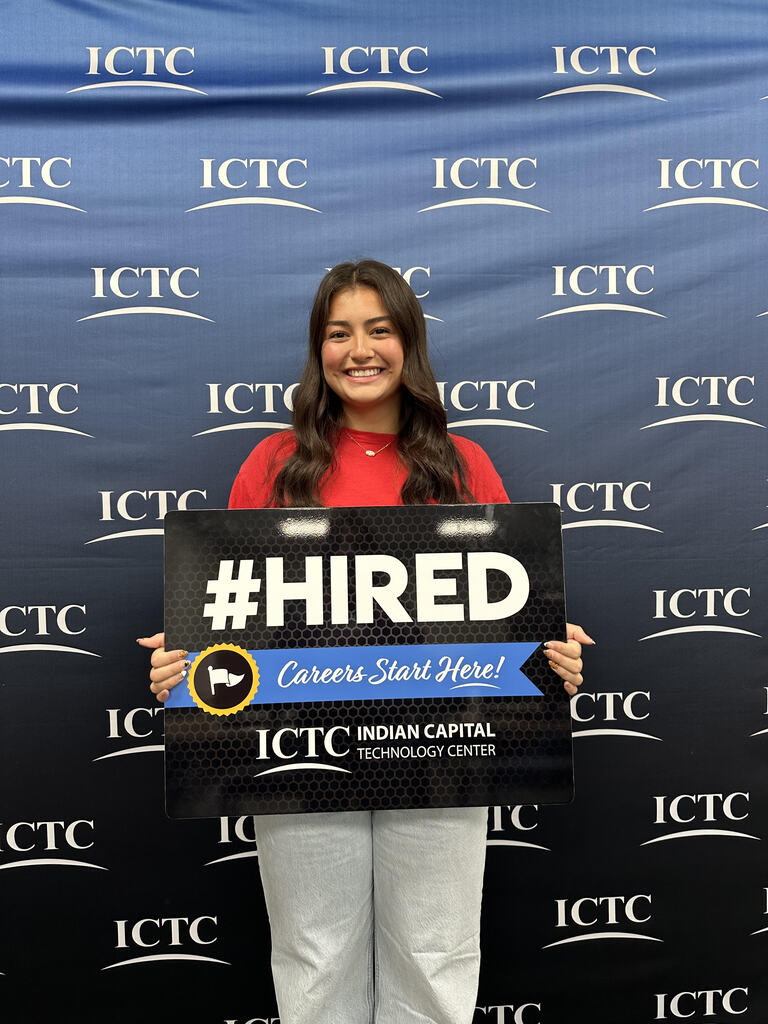 Woman in red top and white pants holding a black sign that says "HIRED" with "Careers Start Here" in front of a blue backdrop.