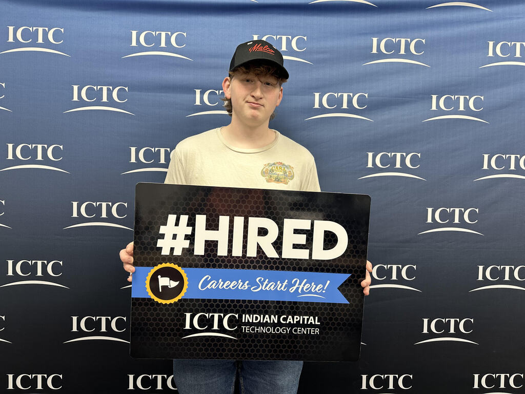 Person in a white shirt holding a sign that says "#HIRED" in front of a blue backdrop.