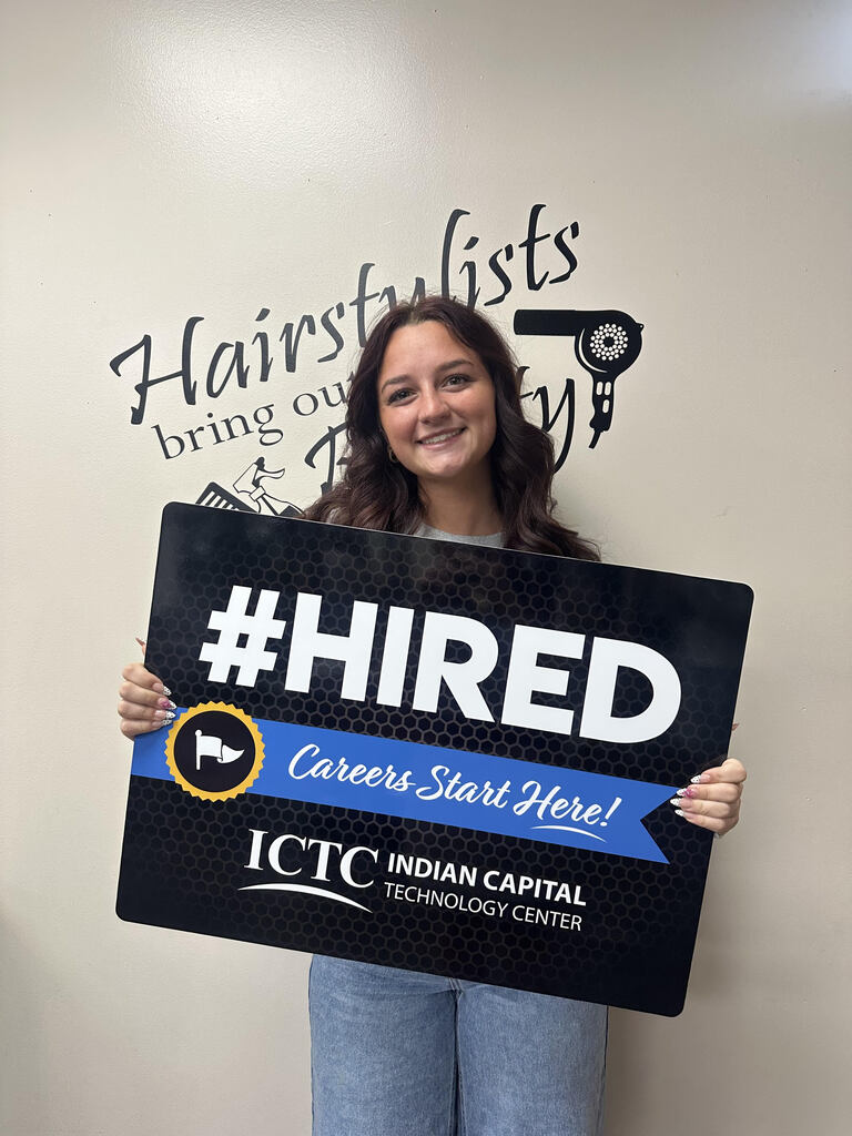 A woman holds a sign that says "#HIRED" in front of a white wall. The sign is blue and black.