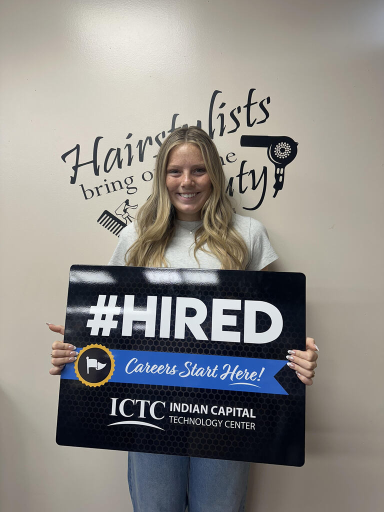 A smiling woman with blonde hair holds a sign reading "#HIRED Careers Start Here!" in front of a wall.