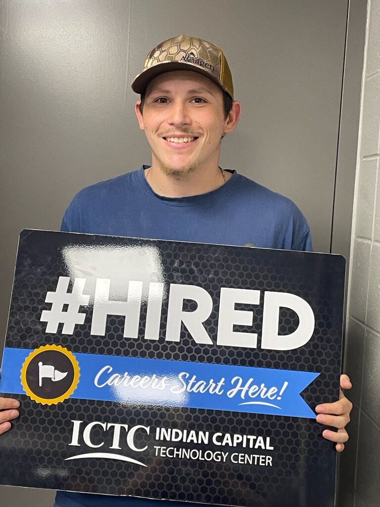 A man with a hat holds a sign that says "#HIRED Careers Start Here!" in front of a gray wall.