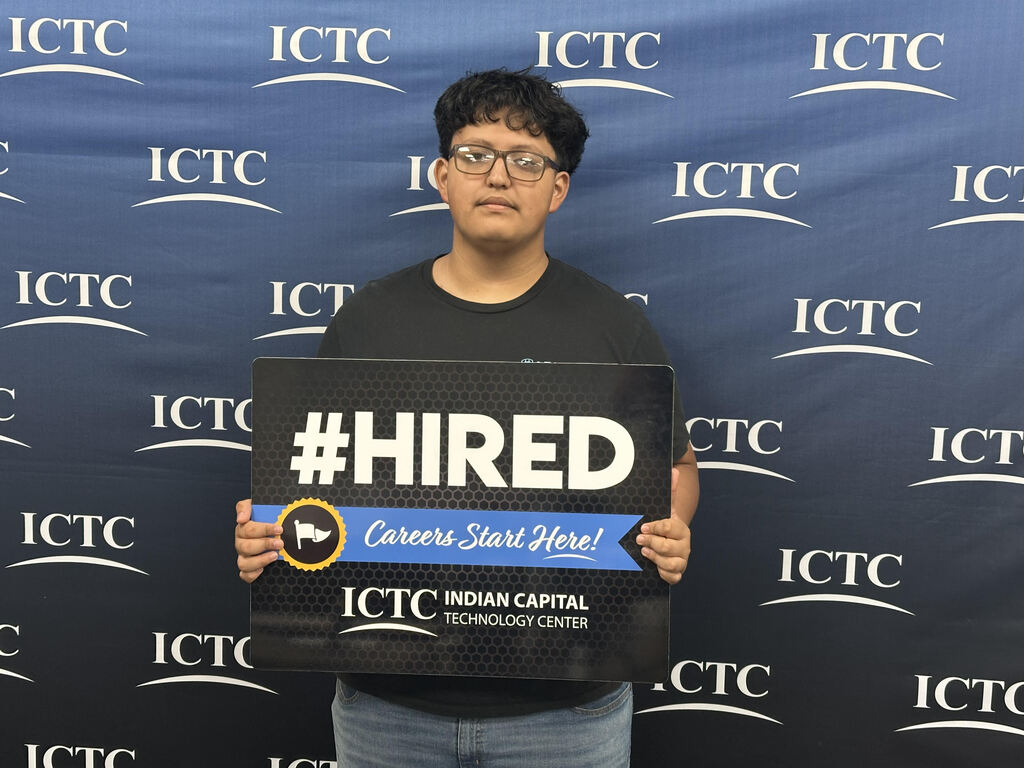 Person in black shirt and glasses holds a sign that reads "#HIRED". They stand in front of a blue backdrop with "ICTC" written repeatedly.