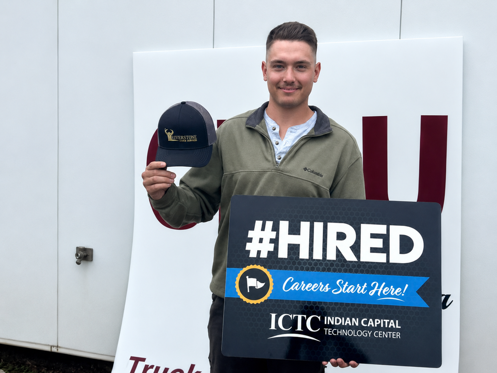 A man wearing a green jacket holds a sign that says "#HIRED" and a cap. The sign has the logo for the Indian Capital Technology Center.