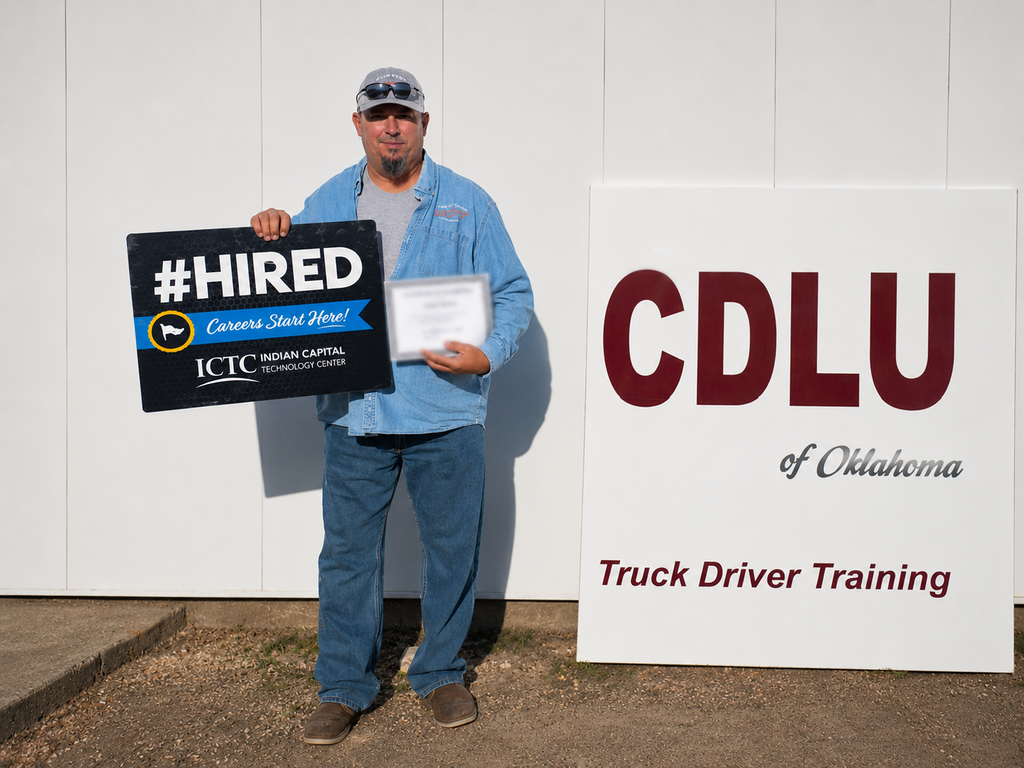 Person holds sign reading "#HIRED" and certificate. Behind them is a sign with "CDLU of Oklahoma" and "Truck Driver Training".