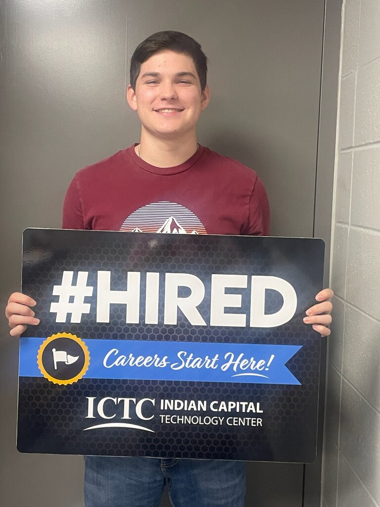 A man holding a black sign with white letters that reads "#HIRED Careers Start Here!" He is smiling.