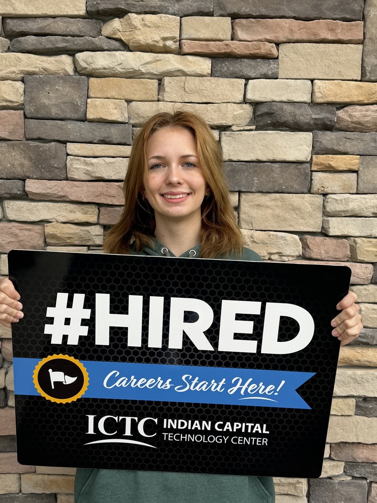 A woman stands with a sign reading "#HIRED Careers Start Here!" in front of a stone wall.