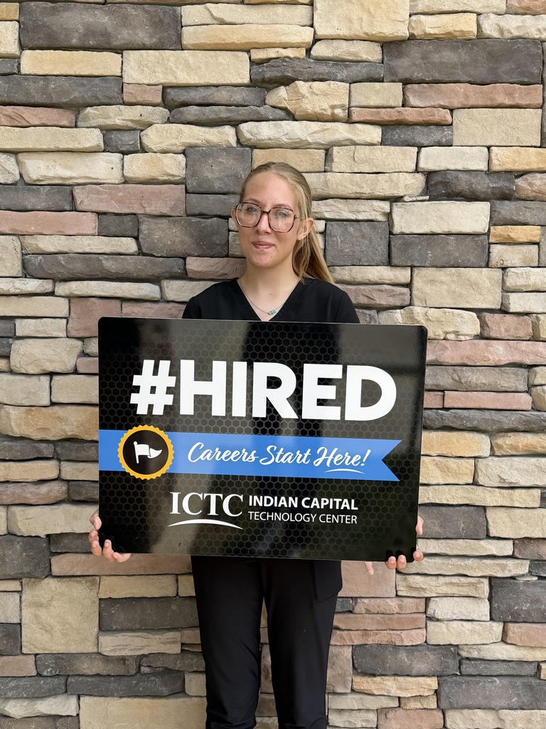A woman with glasses holds a "#HIRED" sign in front of a textured brick wall.