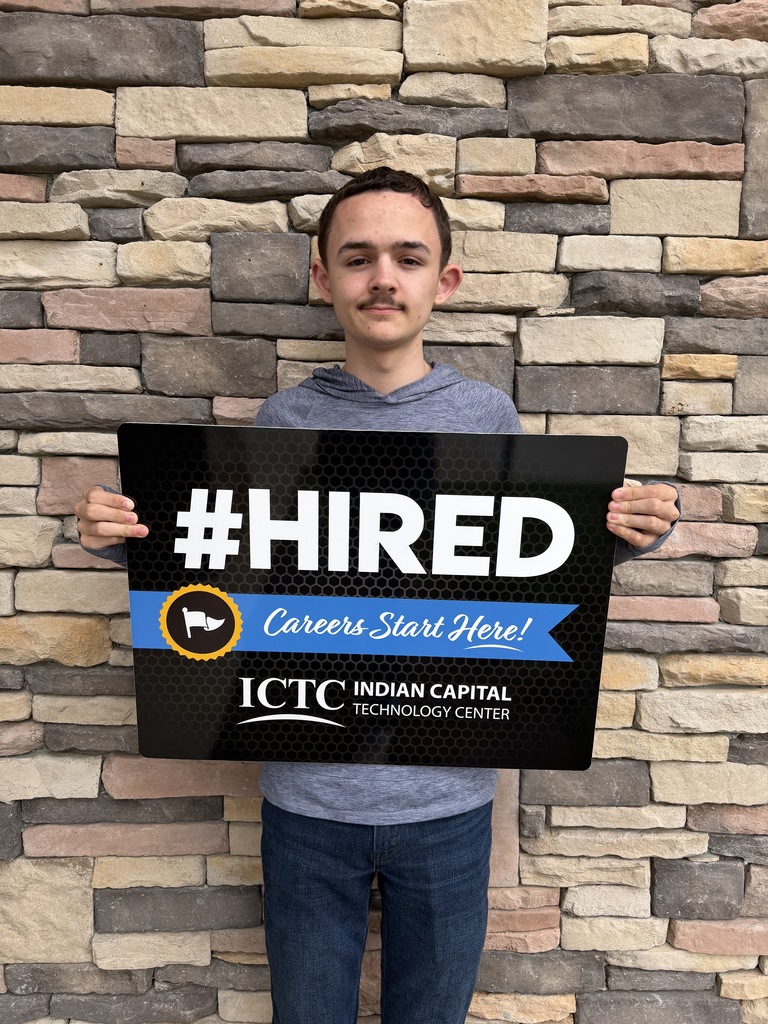 Person stands against brick wall holding a black sign with "#HIRED" in large white letters and a blue banner.