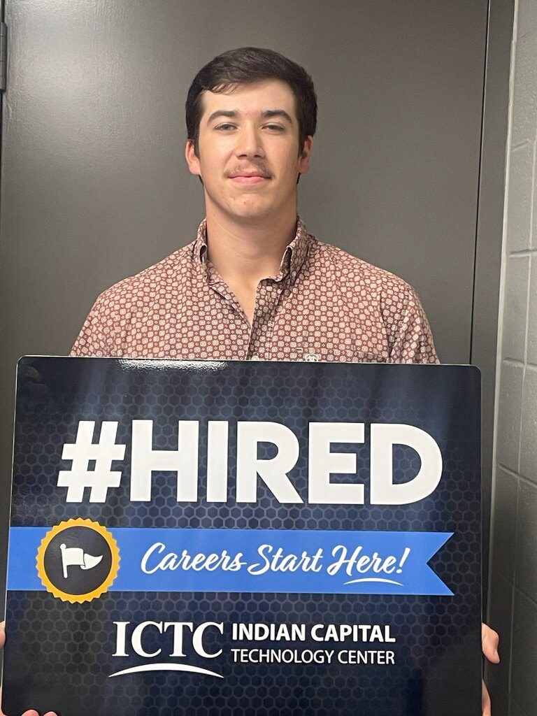 Man standing in an office, smiling, holding a sign that says "#HIRED Careers Start Here!" from the Indian Capital Technology Center.