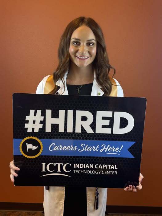 Woman smiling, holding a sign that says "#HIRED Careers Start Here!" against a red wall.