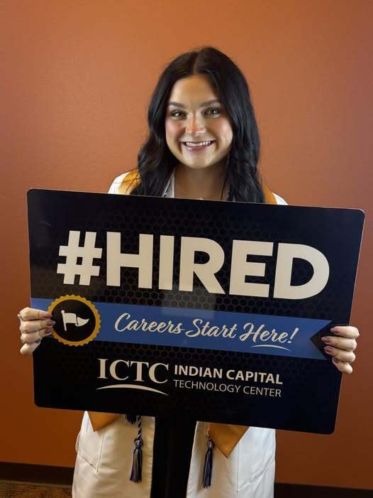 A woman in a graduation gown holds a sign reading "#HIRED" and "Careers Start Here!" in front of a warm background.