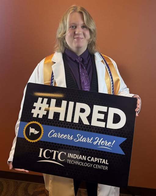 A person in a cap and gown holds a sign saying "#HIRED" and "Careers Start Here!" against a wall.