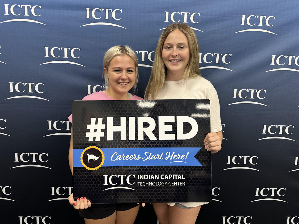 Two women pose with a black sign reading "#HIRED" in front of a blue background with "ICTC" text.