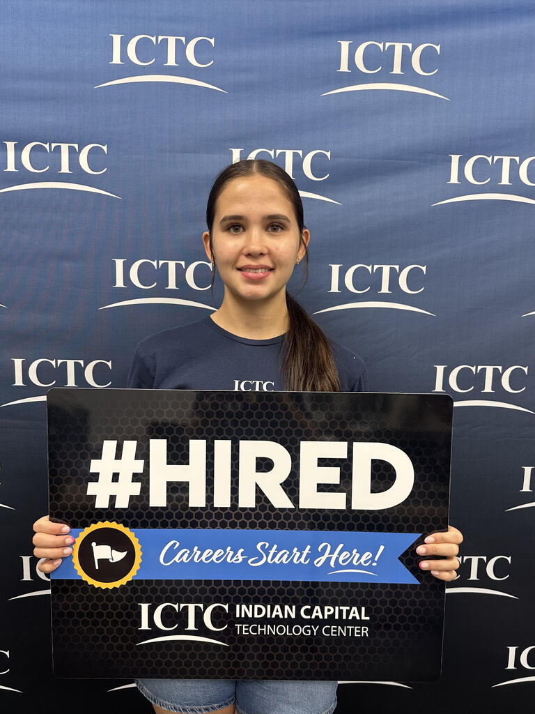 Woman standing against a blue backdrop with multiple ITC logos, holding a sign that says "#HIRED Careers Start Here!"