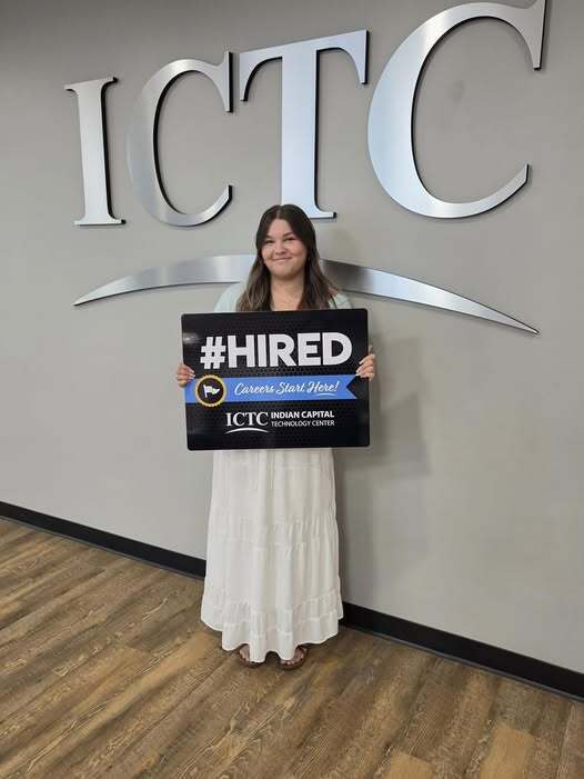 A woman stands before a gray wall with large silver letters reading "ICTC" and holds a sign that reads "#HIRED."