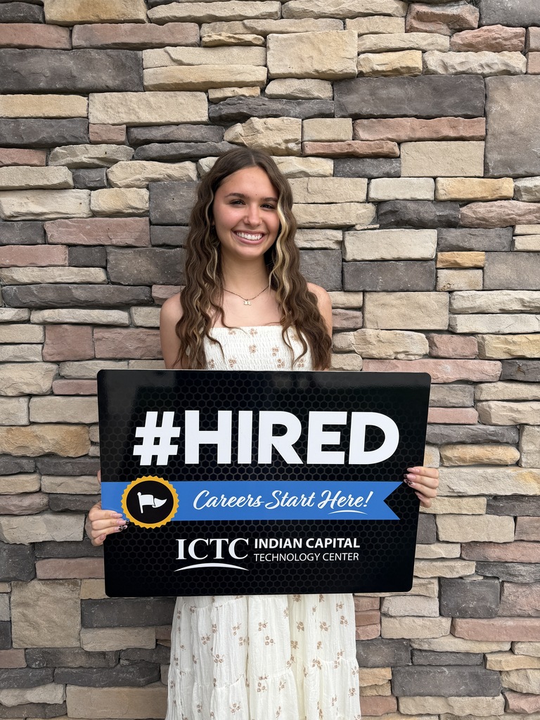 A woman in a white dress holds a black sign reading "#HIRED" in front of a brick wall.