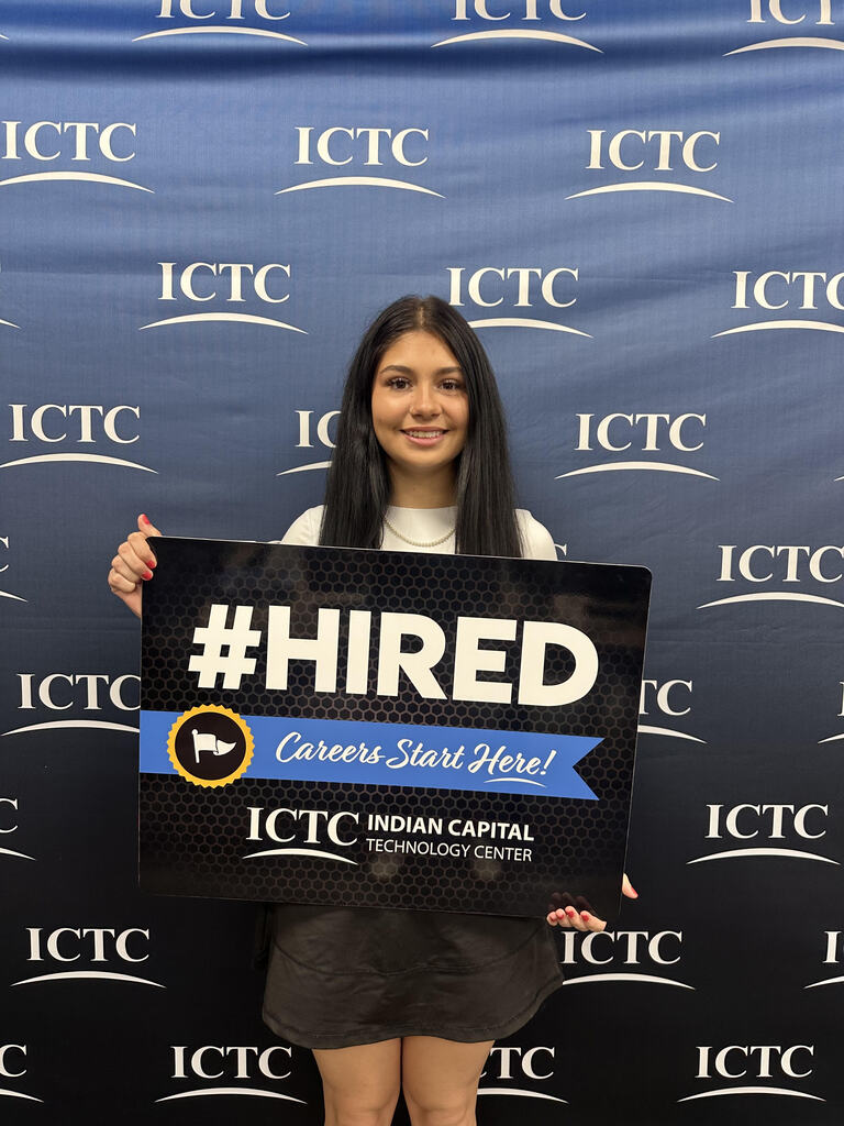 A woman poses with a sign that says "#HIRED" in front of a blue banner with "ICTC" written multiple times.