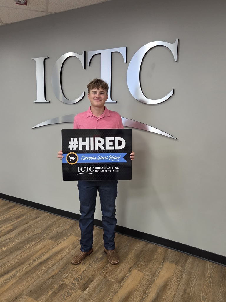 A man in a pink shirt and jeans holds a sign that says "HIRED" in front of an "ICTC" sign.