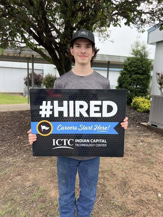 A young man holds a sign reading "#HIRED Careers Start Here!" outside a building with trees and plants.