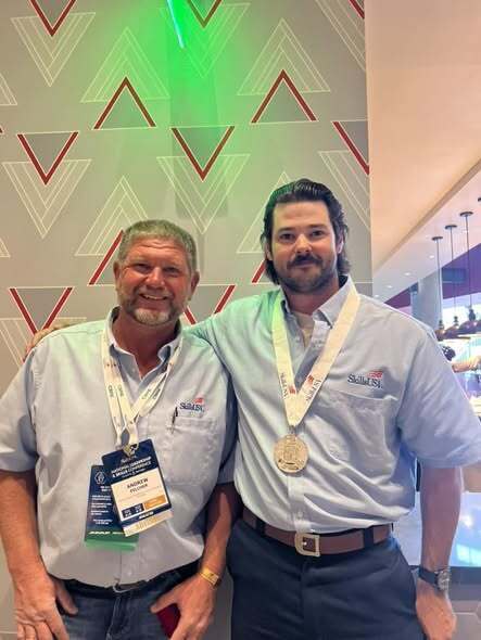 Two men wearing light blue shirts and name tags pose for a photo. One has a silver medal.