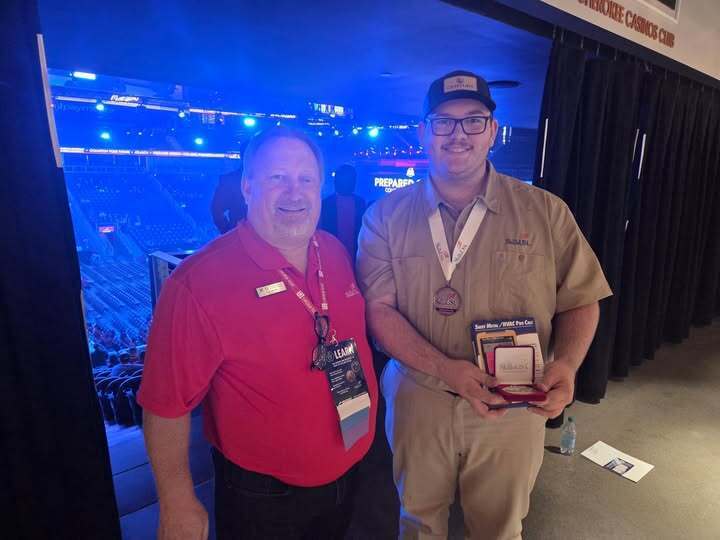 Two men stand in an arena, one in a red shirt, the other in tan, holding an award.