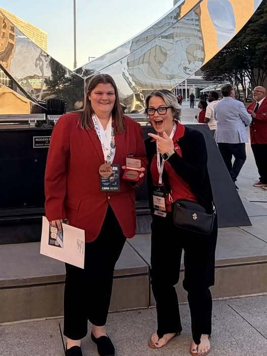 Two women, one holding a paper and a medal, smiling and posing next to a large reflective sculpture.