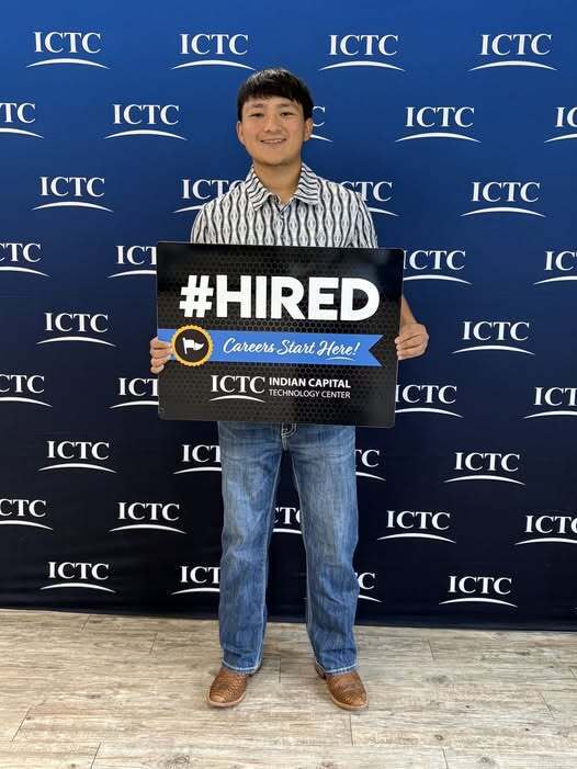 A man in striped shirt and jeans stands before a blue banner holding a sign that reads "#HIRED" in bold letters.