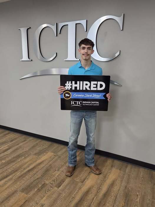 Person in blue shirt and jeans stands in front of ICTC sign, holding a sign that reads "HIRED".