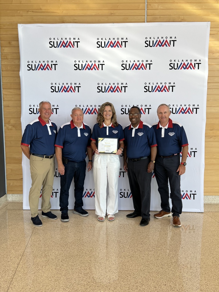 Five adults in blue shirts and white pants stand in front of a white backdrop with red and blue text.
