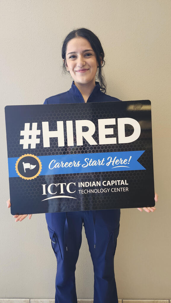 A person in blue holds a black sign that reads "#HIRED Careers Start Here!" in front of a plain wall.