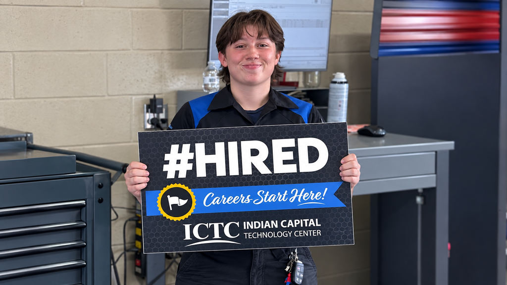 A young person holds a #HIRED sign in a workshop with a gray desk, computer, and tools.